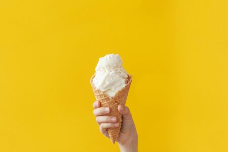Children's hand holds delicious vanilla ice cream cone on a bright yellow background with copy space.の写真素材
