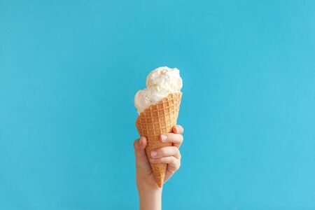 Children's hand holds delicious vanilla ice cream cone on a bluebackground with copy space.の写真素材