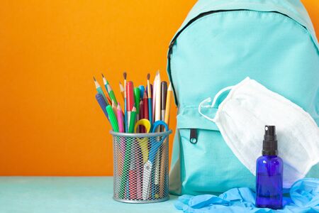 Blue School backpack with mask, hand sanitizer, gloves and stationery on table on orange background....