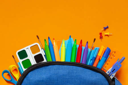 Full blue School Backpack with different supplies on orange background. FLat lay. Concept back to...