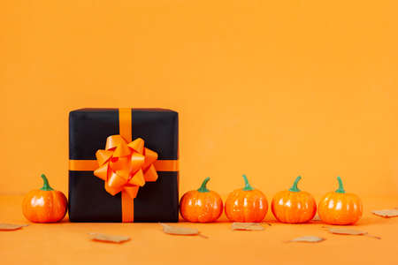 Black present box with orange ribbon and pumpkin on orange background. Happy Halloween concept.