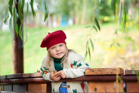 Beautiful smiling blonde girl in a burgundy beret and knitted sweater in sunny warm autumn day. Child playing in park at autumn.の写真素材