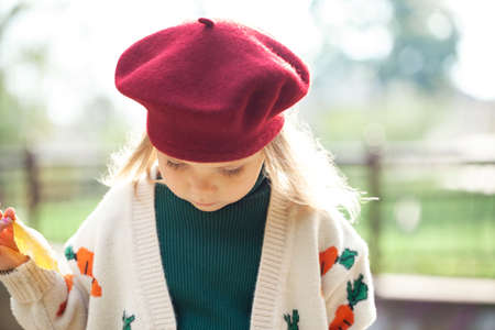 Cute little blond girl wearing burgundy beret and knitted sweater in autumn park, close-up.の写真素材