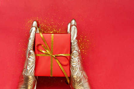 Hands in golden medical gloves holding Christmas red gift box with golden bow on red background....