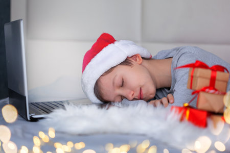 Cute little boy in Santa hat and Christmas pajamas fell asleep near laptop while waiting for christmas presents at home. New Year and Holidays concept.の写真素材