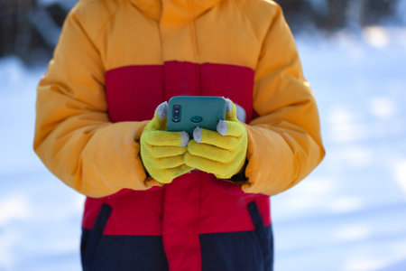 Hands in wool yellow gloved holds smartphone on the background of the snowy winter forest. Touch Screen Gloves.の写真素材