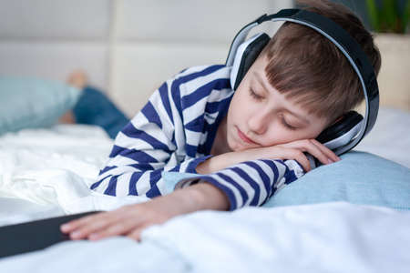 Beautiful child boy lies on the bed and listens to music or a podcast and enjoys. Child got tired of playing the phone and fell asleepの写真素材