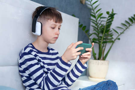 Child Boy in headphone looking and playing phone, learning on the bed in living room. Concept kids using technologyの写真素材