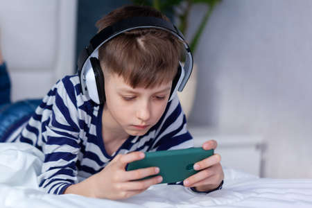 Child Boy in headphone looking and playing phone, learning on the bed in living room. Concept kids...