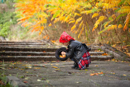 Cute girl child in black jacket collects autumn leaves in the hat. Girl 4 years old walks in the autumn park. Happy kid.の写真素材