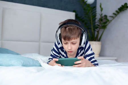 Child Boy in headphone looking and playing phone, learning on the bed in living room. Concept kids using technologyの写真素材