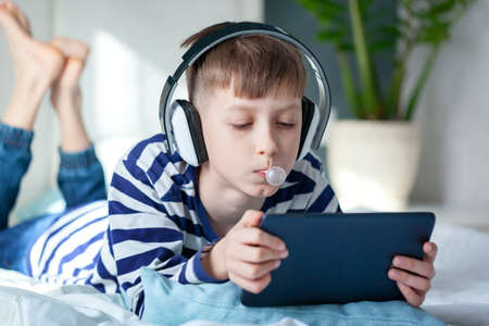 Cute child boy in headphone playing on a digital tablet on the bed in living room. Education, free...