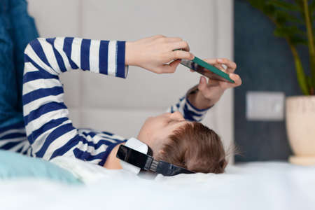Child Boy in headphone looking and playing phone, learning on the bed in living room. Concept kids using technologyの写真素材