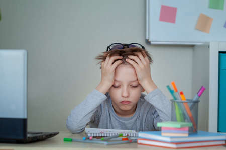 Unhappy and strssed school boy studying with a notebooks on his desk at home. Child is tired of online learning. Education and childhood conceptの写真素材