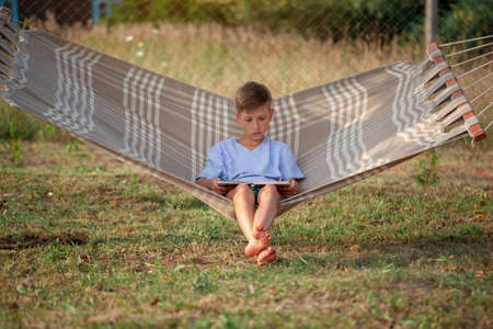 Cute child boy reading book and dreamink on hammock in backyard on summer day. Kid swinging and relaxing in hammock.の写真素材