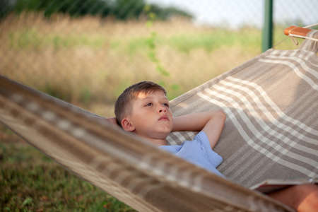 Cute child boy reading book and dreamink on hammock in backyard on summer day. Kid swinging and relaxing in hammock.の写真素材