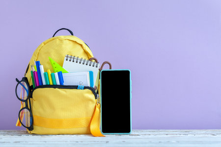 concept back to school. Full yellow school backpack and smartphone with stationery on table on purple background.の写真素材
