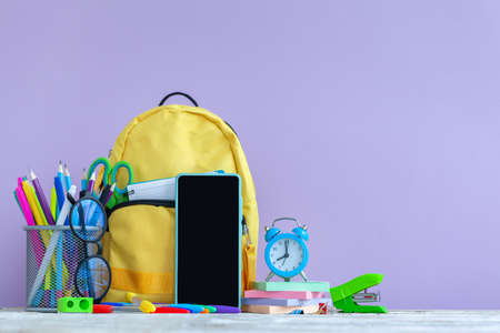 concept back to school. Full yellow school backpack and smartphone, eyeglasses, alarm clock with stationery on table on purple background.の写真素材