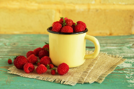 A metal cup with strawberries and raspberries on the napkins from sackcloth on the blue-white wooden table on the background of the brick wall.の写真素材