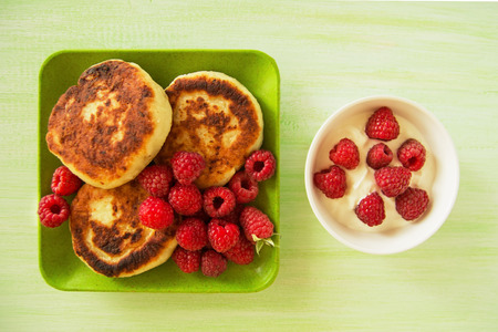 Cottage cheese pancakes with fresh raspberries and sour cream on the green and white plates on the green wooden background, top view.の写真素材
