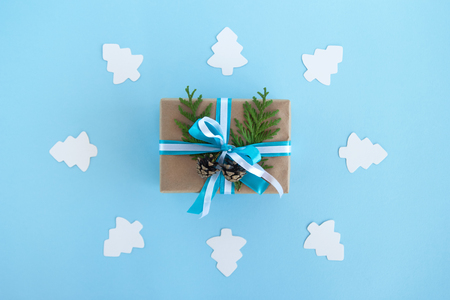 Gift box wrapped of craft paper, blue and white ribbons and decorated fir branches and pinecones on the blue background with white paper fir tree, top view. Christmas present.の写真素材