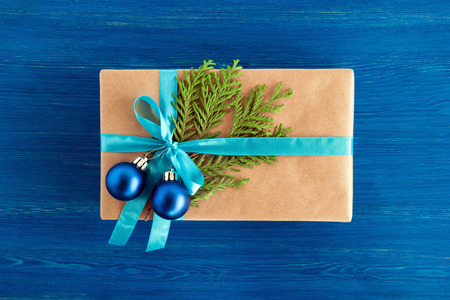 Gift box wrapped of craft paper, blue ribbon and decorated fir branches and blue Christmas balls on the blue wooden background, top view. Christmas present.の写真素材