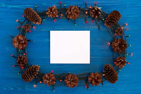 Top view on frame from Christmas lights, pine cones and white sheet of paper on the blue wooden background with copy space.の写真素材