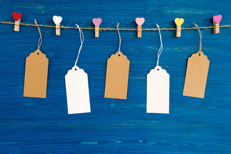 Brown and white blank paper price tags or labels set and wooden pins decorated on colored hearts hanging on a rope on the blue wooden background.の写真素材