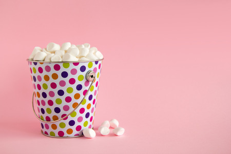 Small colorful bucket filled with white marshmallows on a pink pastel background. Minimal concept with copy space.の写真素材