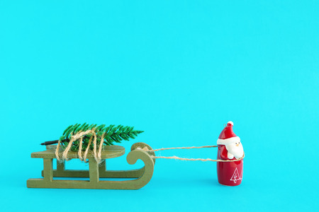 Santa Claus is carrying a Christmas tree on a green wooden sleigh on a bright blue background....