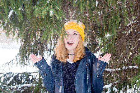 Young smiling woman with blue eyes and blond hair in a yellow knitting hat and black leather jacket under the fir tree at the moment of snowfall.の写真素材