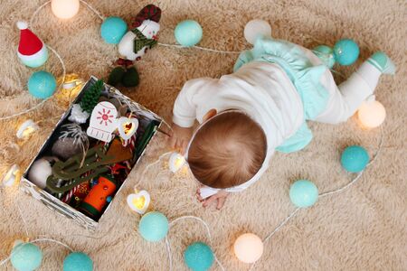 Little girl is playing with blue and white Christmas lights and box with Christmas decorations on a...