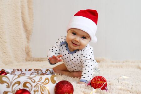 Adorable smiling little girl in Santa Claus red hat is playing with red shiny Christmas decorations...