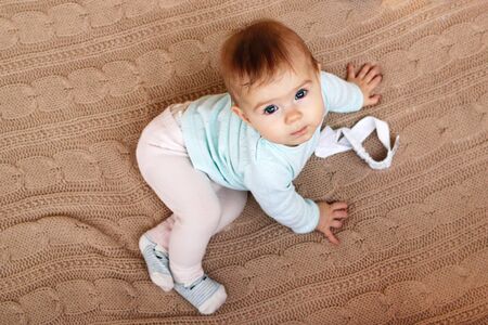 Beautiful little girl with blue eyes and blondie hair is sitting on a beige knitted plaid and...