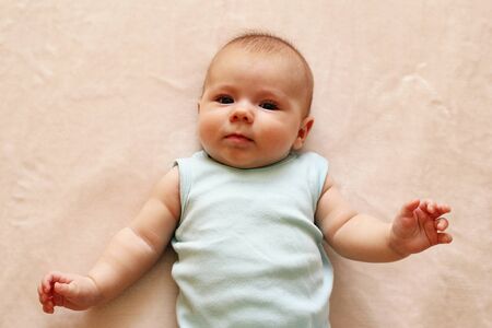 Cute three month baby in blue bodysuit is lying on a beige blanket and looking at the camera.