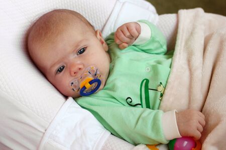 Cute infant little girl with pacifier is lying in her bed under beige plaid.