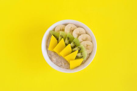 Bowl of oatmeal with banana, kiwi and mango on yellow background. Flat lay with copy space.の写真素材