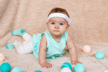 Cute little girl with beautiful blue eyes in blue dress is sitting on a beige plush plaid with Christmas lights.の写真素材