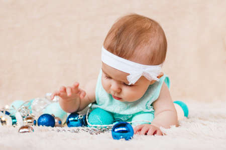 Cute little girl in blue and white dress is playing with Christmas decorations on a beige plush plaid with Christmas lights.の写真素材