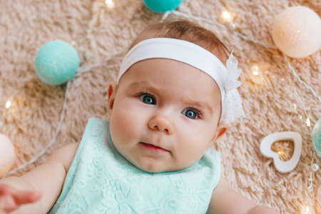 Portrait of cute little girl with beautiful blue eyes in blue dress on a beige plush plaid with Christmas lights, top view.の写真素材