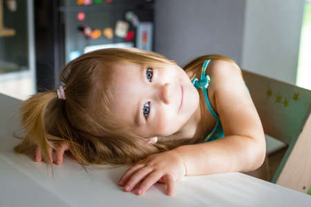 Adorable baby girl with blond hair and blue eyes with hand on table. Sunny morning at home.の写真素材