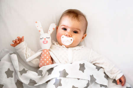 Candid portrait of cute Caucasian baby girl six months old lying on white bed with pacifier and toy of hare.の写真素材