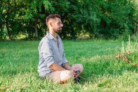 Portrait of young handsome European man in casual clothing sitting on a grass in summer park.の写真素材