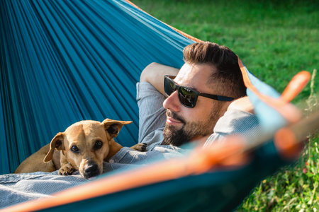 Smiling attractive European man in sunglasses is resting in hammock with his cute little dog.の写真素材