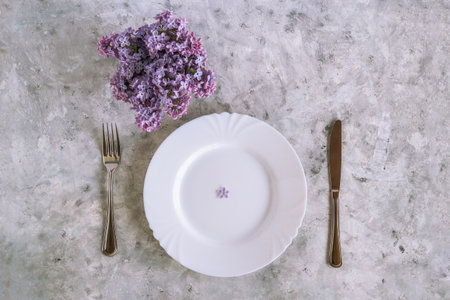 Served plate with purple lilac flower with five petals, fork and knife, bouquet of lilac flowers on gray concrete background, top view.の写真素材