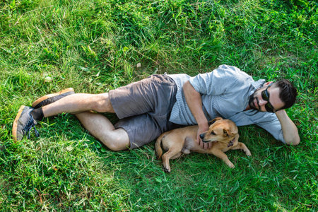 Attractive European man in casual clothing and sunglasses is lying on grass and hugging his cute little dog.の写真素材