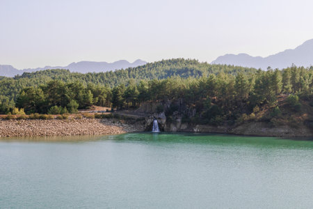 Tranquil Forest Lake with Rocky Waterfall and Mountainous Horizonの写真素材