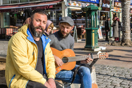 Istanbul, Turkey - December 29, 2022: Two men are sitting on bench by the street and one of them playing guitar.のeditorial素材