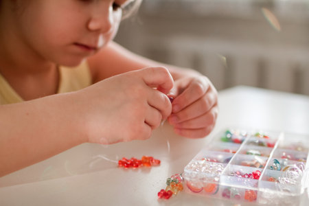 A young girl with pigtails creates colorful jewelry by stringing beads at a well-lit table, surrounded by an assortment of beads and string, showing her joy and focus in crafting.の写真素材