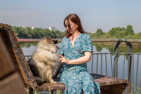 Thoughtful woman in a blue dress taking a break with her Keeshond dog by the lake on a sunny day.